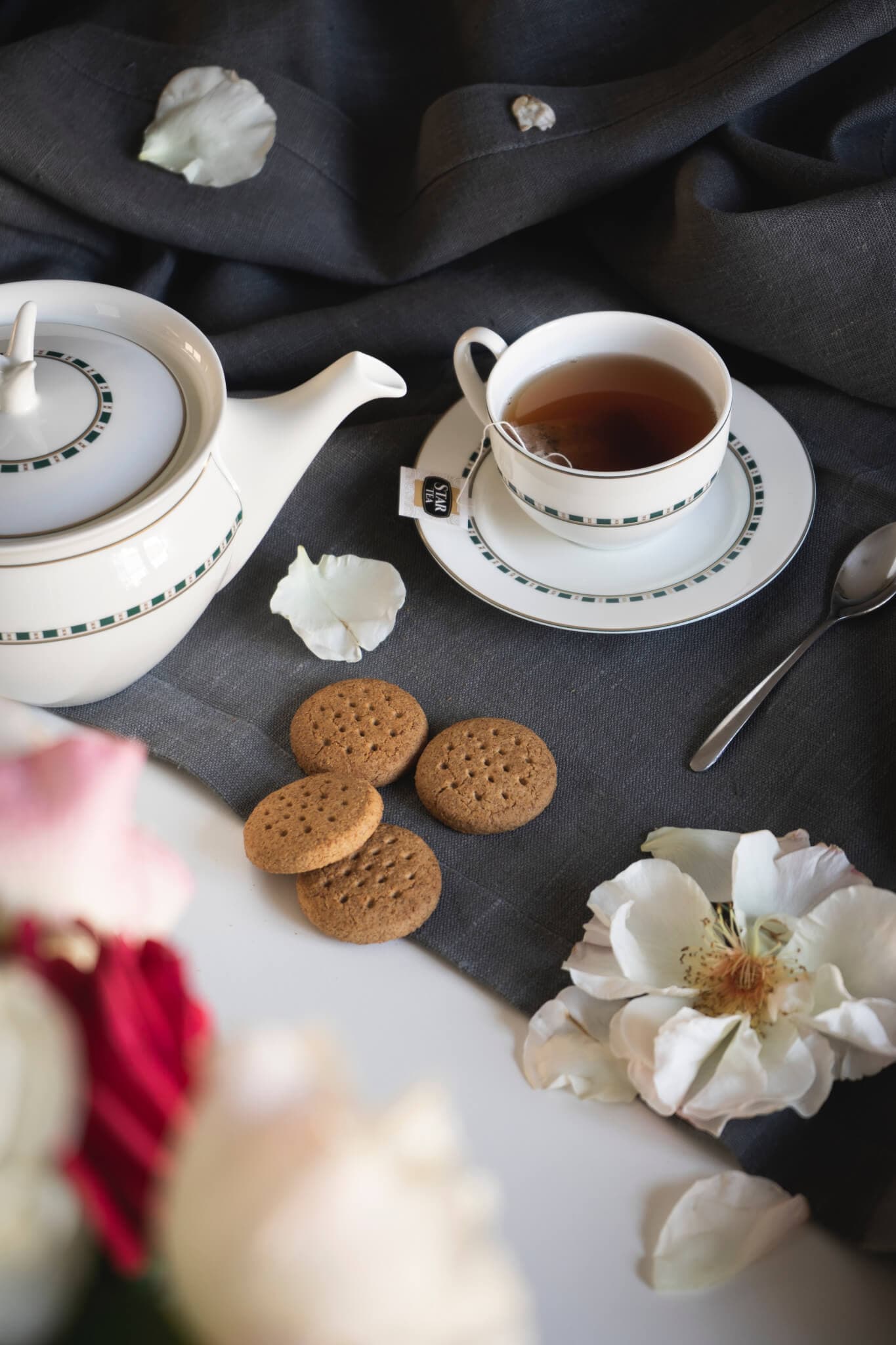 Teapot, teacup with tea, cookies, spoon, and flower petals on a gray tablecloth beside a bouquet of flowers. - Home Instead