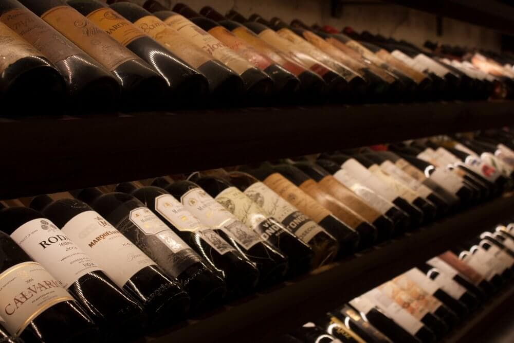 Rows of wine bottles arranged on shelves with various labels, in a dimly lit setting. - Home Instead