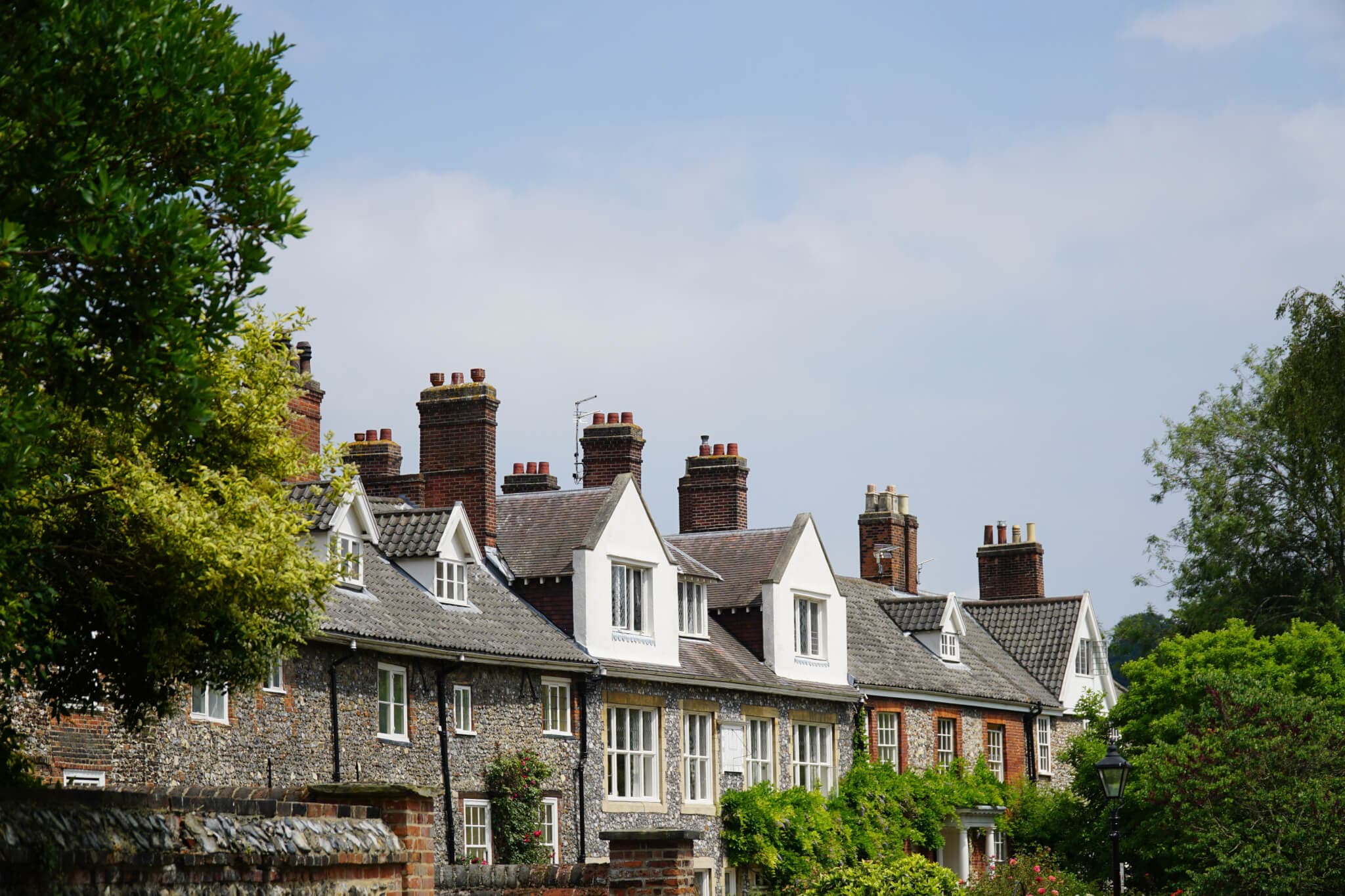 Row of picturesque, traditional English cottages with brick chimneys, surrounded by lush greenery under a blue sky. - Home Instead