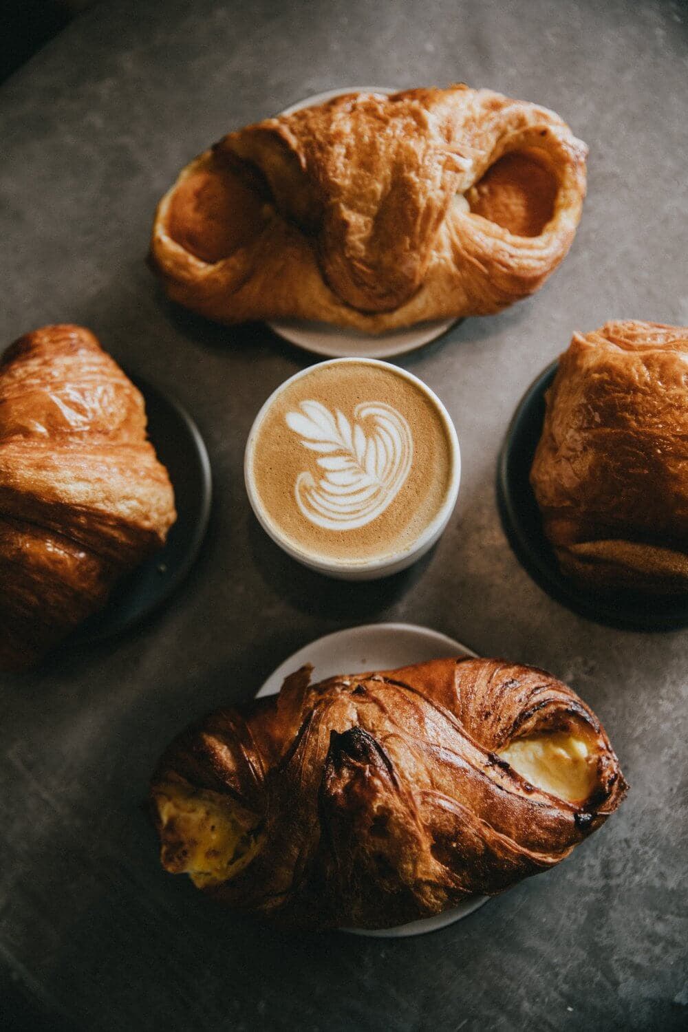 Four pastries surround a latte with a leaf design in the foam, all set on a dark table. - Home Instead