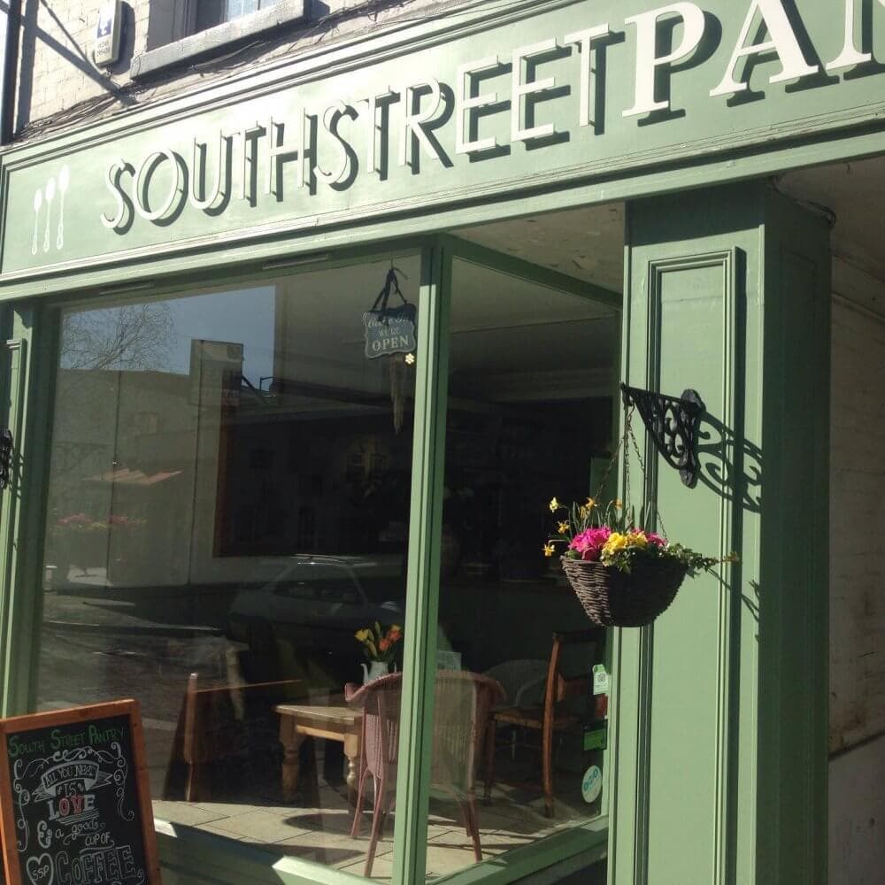 A cozy café with green exterior named "Southstreet Pantry," featuring a hanging flower basket and chalkboard sign outside. - Home Instead