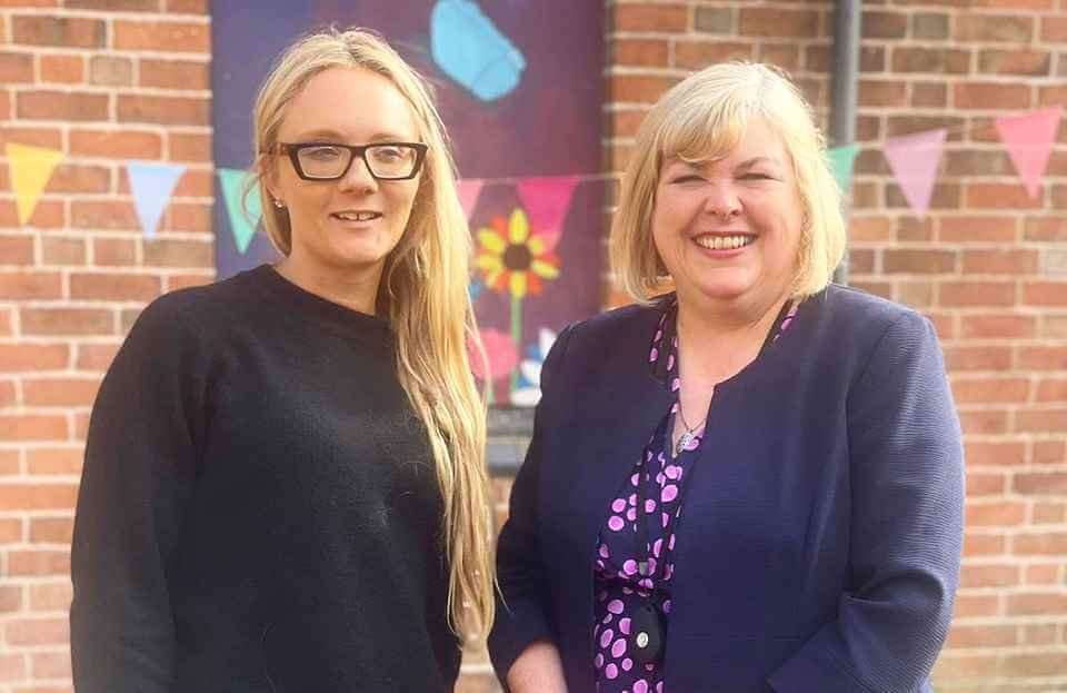 Two women standing together and smiling in front of a brick wall with colorful bunting in the background. - Home Instead