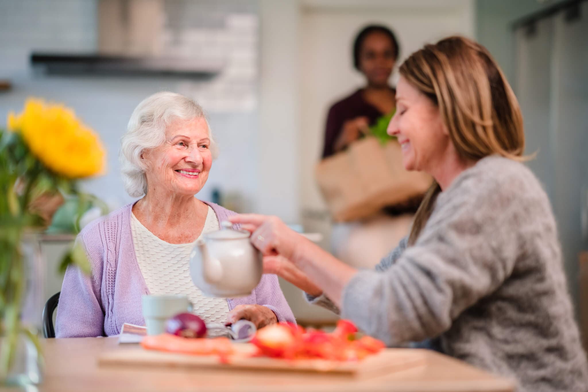 Older woman smiling at younger woman pouring tea, with a third person holding groceries in the background. - Home Instead