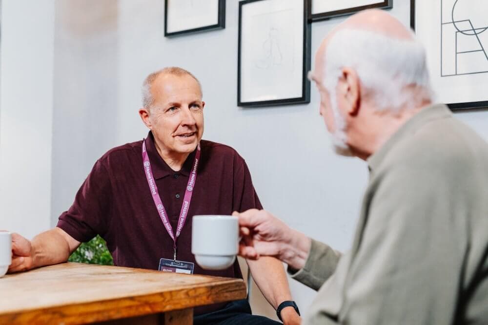 Two men are sitting at a wooden table, having a conversation while holding coffee cups. - Home Instead