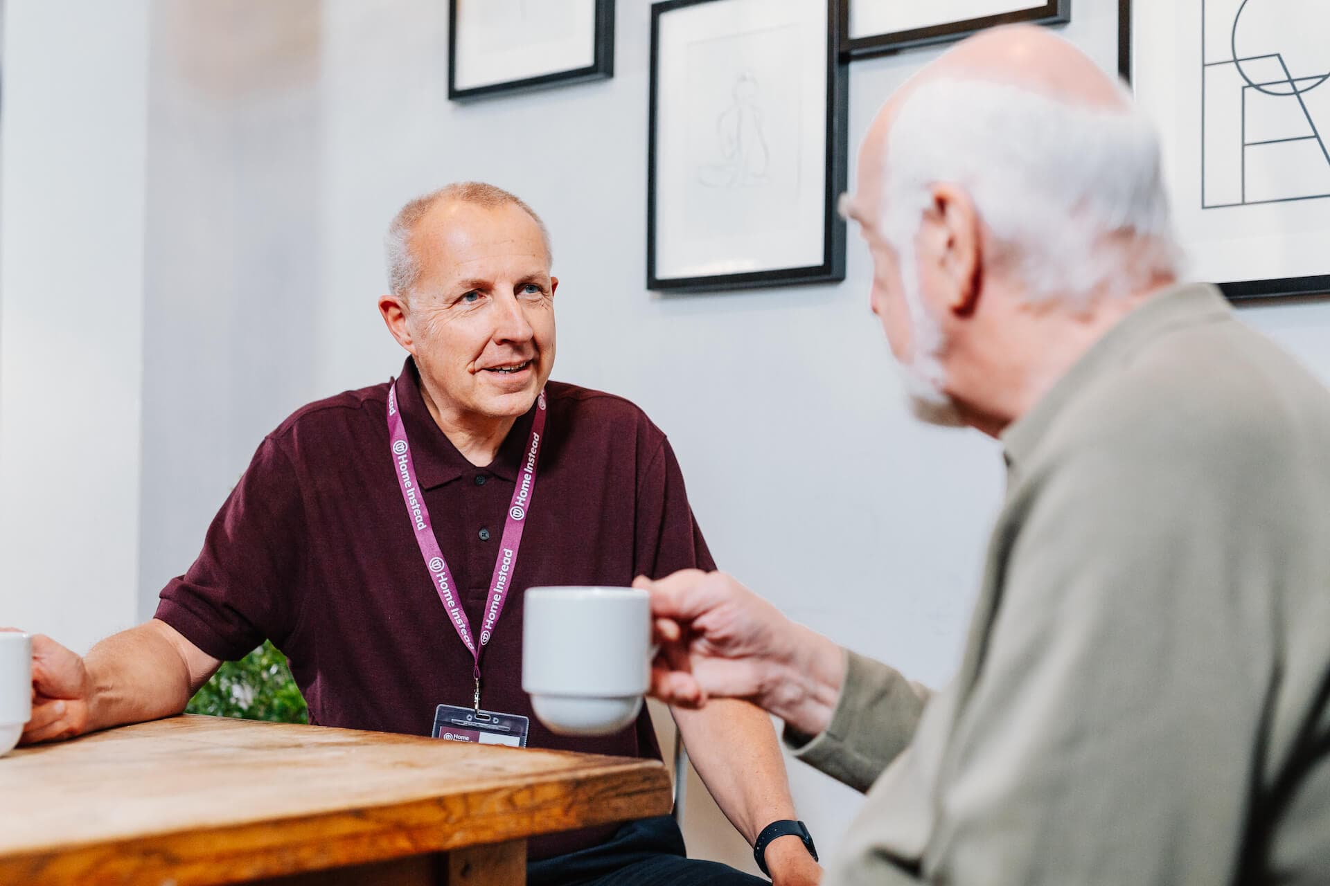 Two men are sitting at a wooden table, having a conversation while holding coffee cups. - Home Instead