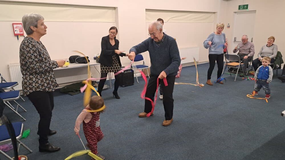 Adults and children enjoying a ribbon dance activity in a community room, engaging and smiling together. - Home Instead
