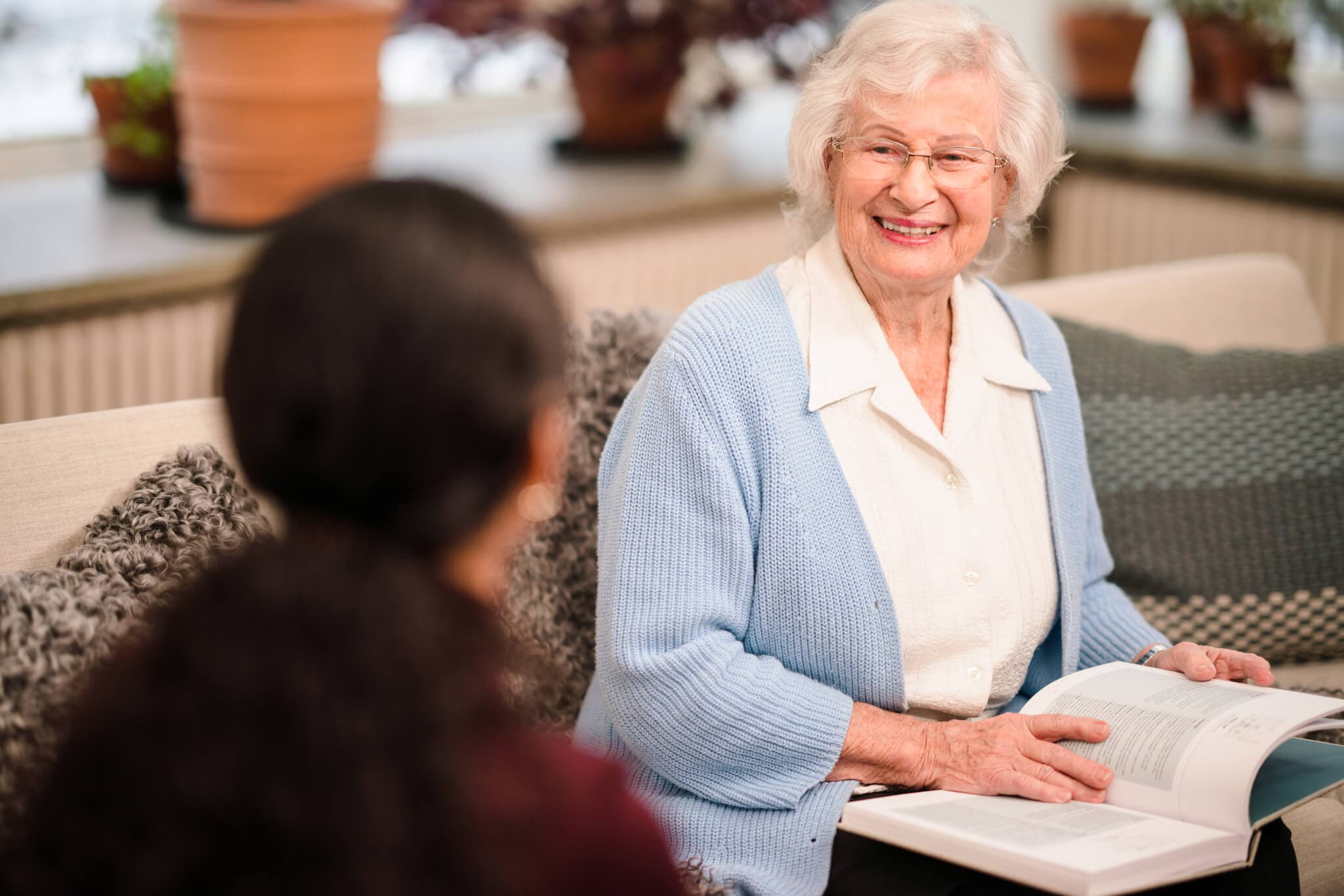 An elderly woman with glasses reading a book, smiling at a person sitting next to her in a cozy room. - Home Instead