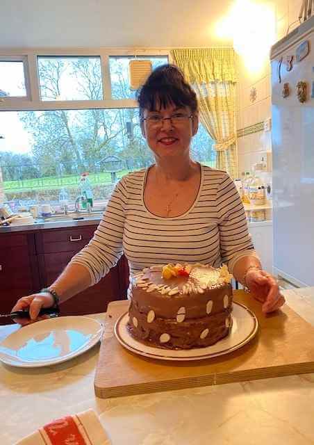A woman in a striped shirt smiles while holding a knife next to a decorated chocolate cake in a cozy kitchen. - Home Instead