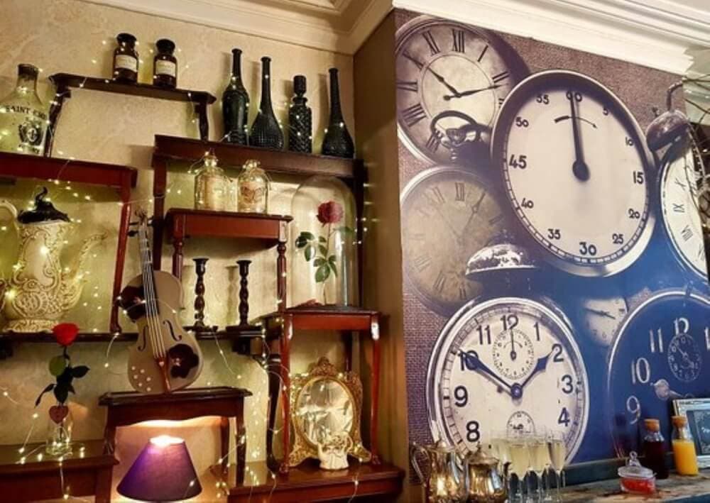 A wall with vintage clocks, shelves with antique bottles, vases, a guitar, fairy lights, and a small lamp. - Home Instead