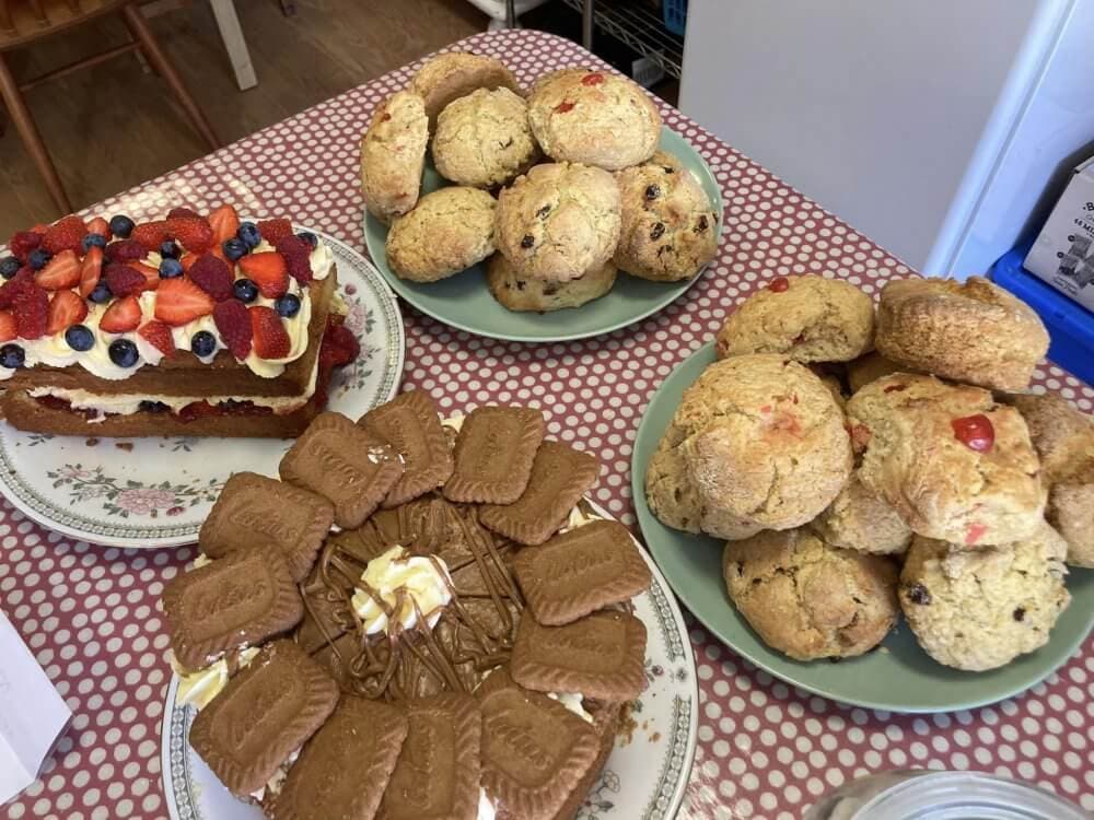 A table with various desserts: a berry-topped cake, Biscoff cake, and plates of scones. - Home Instead