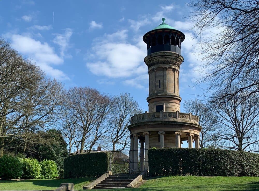A historic stone tower with an observation deck, surrounded by trimmed hedges and leafless trees, under a blue sky. - Home Instead