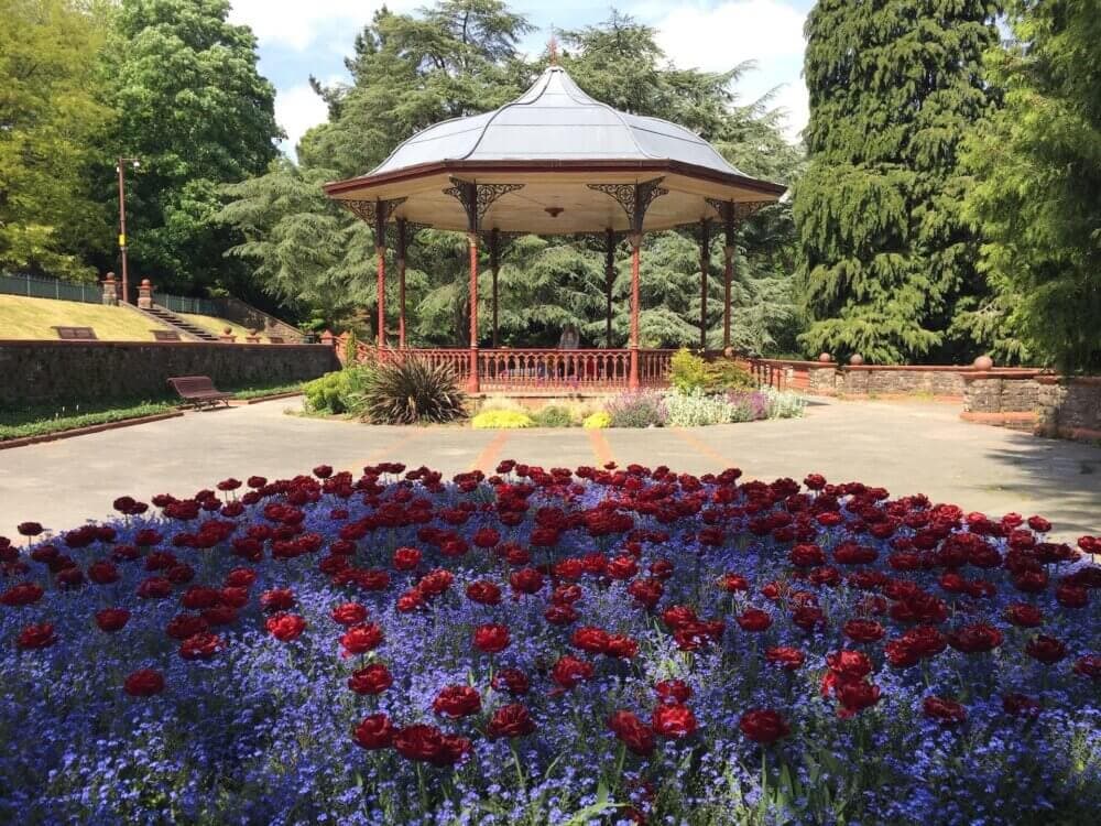 Ornate gazebo in a park with a flower bed of red and blue flowers in the foreground, surrounded by lush green trees. - Home Instead