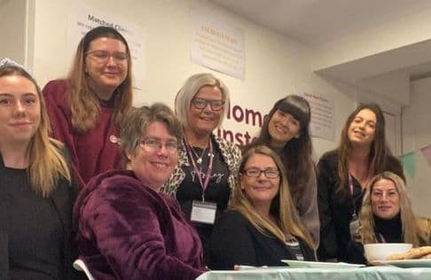 A group of eight women smiling at the camera while seated and standing around a table in a room with posters on the wall. - Home Instead