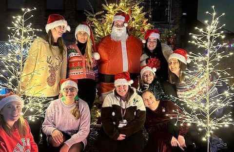 A group of people in Christmas attire, including Santa, pose in front of a decorated tree with festive lights. - Home Instead