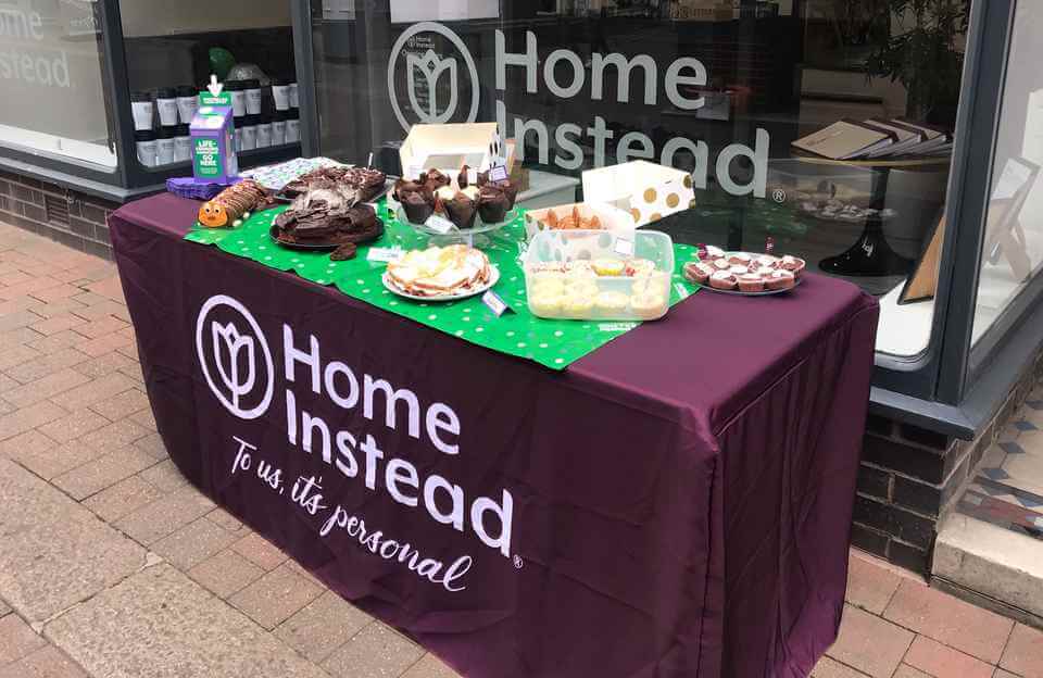 A table with various desserts is set up outside a storefront with a "Home Instead" sign in the window. - Home Instead