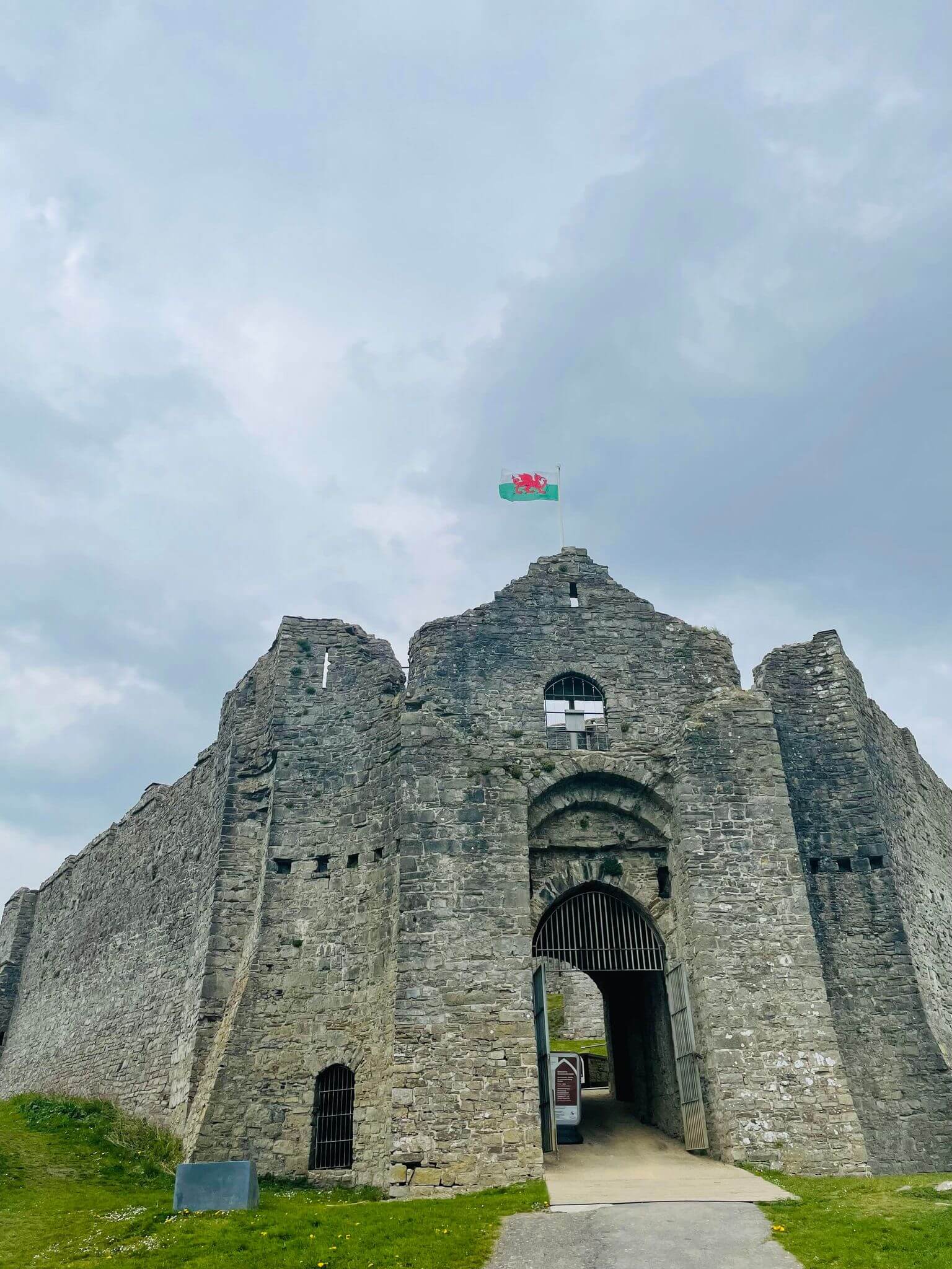 Old stone castle with arched entrance, and Welsh flag flying atop against a cloudy sky. Green grass in the foreground. - Home Instead
