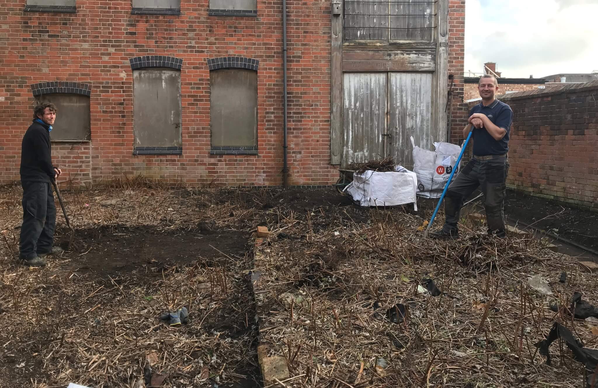 Two people clearing debris in a neglected, overgrown yard of a brick building with boarded-up windows. - Home Instead