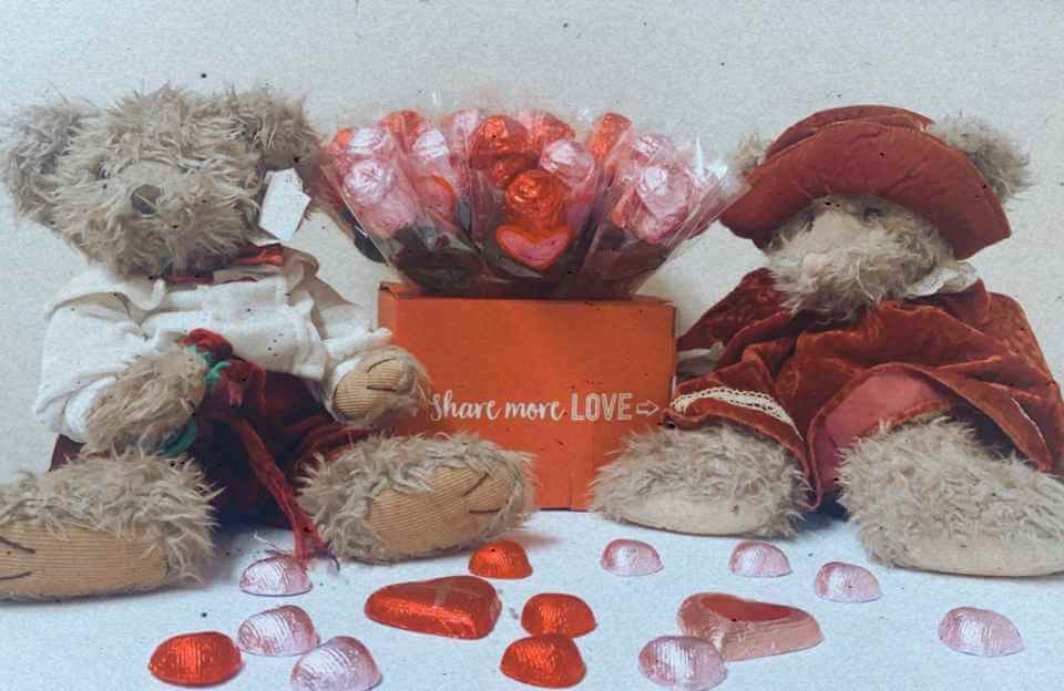 Two teddy bears with heart-shaped chocolates and a "Share More Love" box between them, against a plain background. - Home Instead