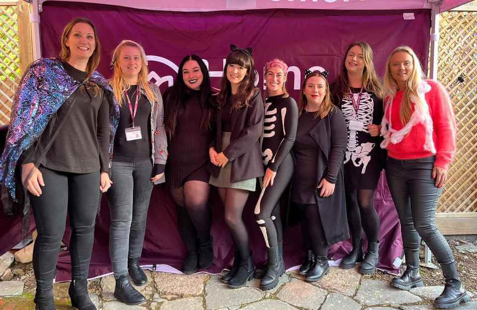 A group of eight people in various costumes pose together in front of a purple tent. - Home Instead