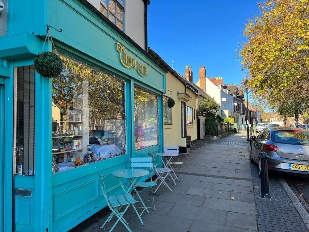 Street view of a teal-themed cafe storefront with outdoor seating, under a bright blue sky and trees lined with autumn leaves. - Home Instead