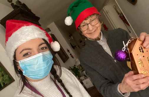 Two people wearing Santa hats, one in a face mask, smiling and holding a gingerbread house and an ornament. - Home Instead