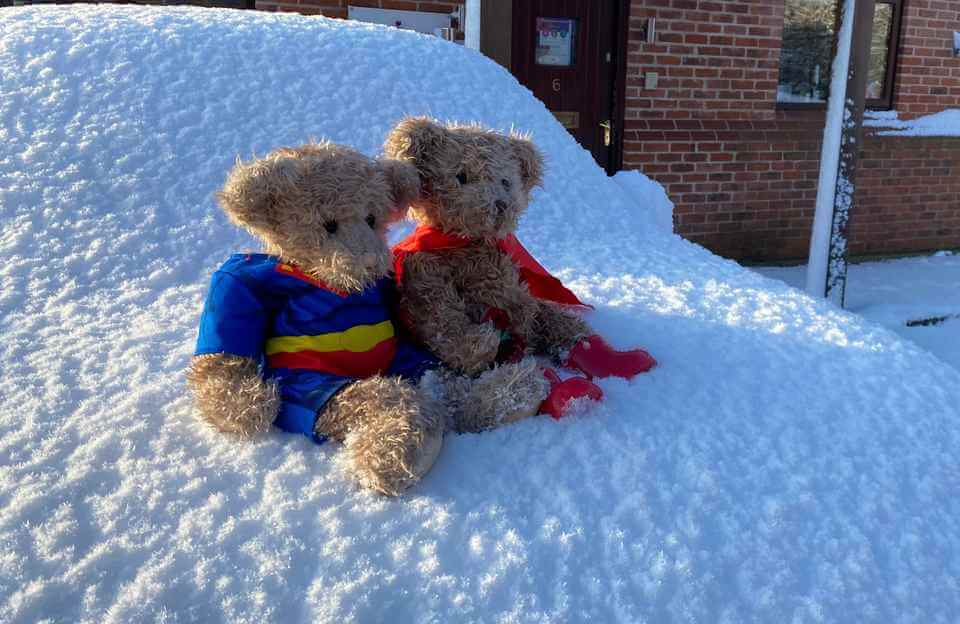Two teddy bears dressed as superheroes sit on a snow-covered car outside a brick building. - Home Instead