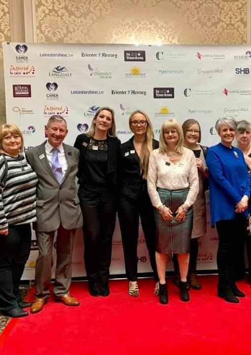 A group of nine people posing together on a red carpet in front of a step-and-repeat banner. - Home Instead