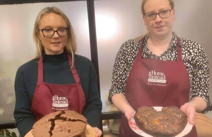 Two women wearing aprons hold homemade cakes; one holds a chocolate cake, and the other holds a chocolate heart cake. - Home Instead
