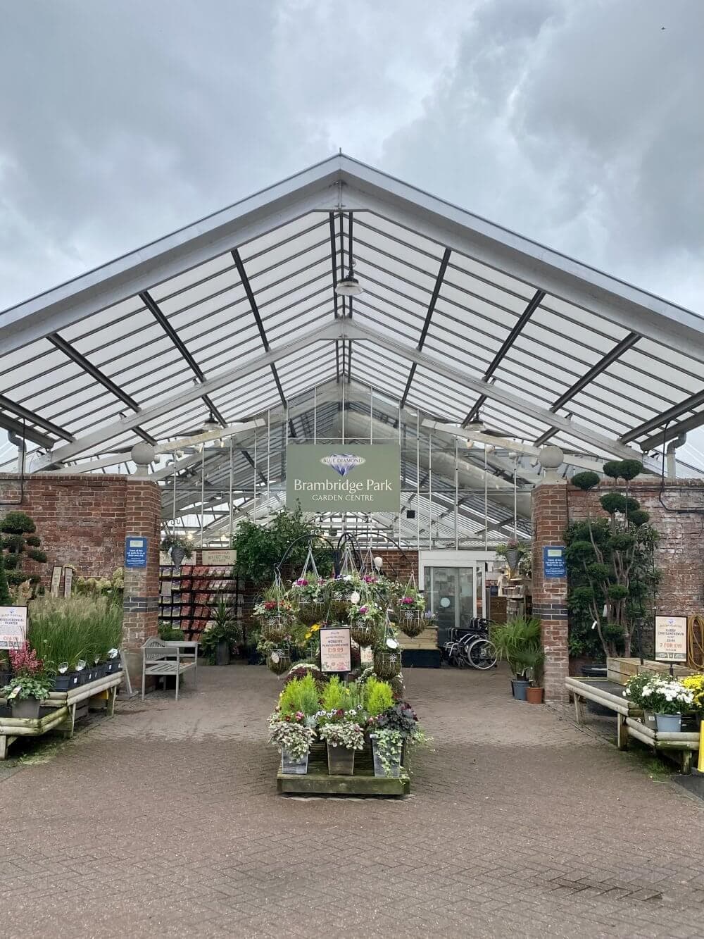 Entrance of Brambridge Park garden centre with various plants and flowers displayed outside under a large glass canopy. - Home Instead