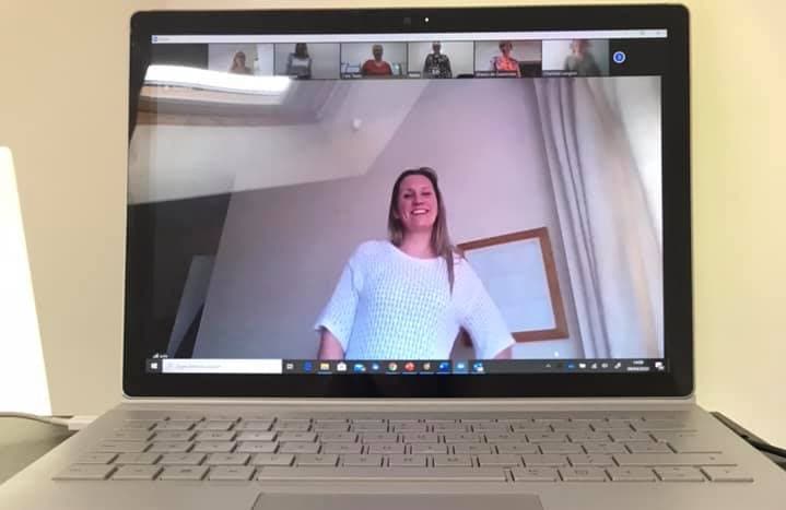 A laptop screen showing a woman standing and smiling during a video conference call with others visible in the top row. - Home Instead