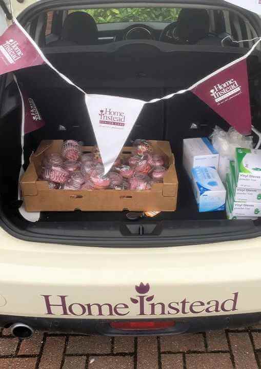 Car boot with "Home Instead" banner, filled with cupcakes, tissue boxes, and face masks on a brick pavement. - Home Instead