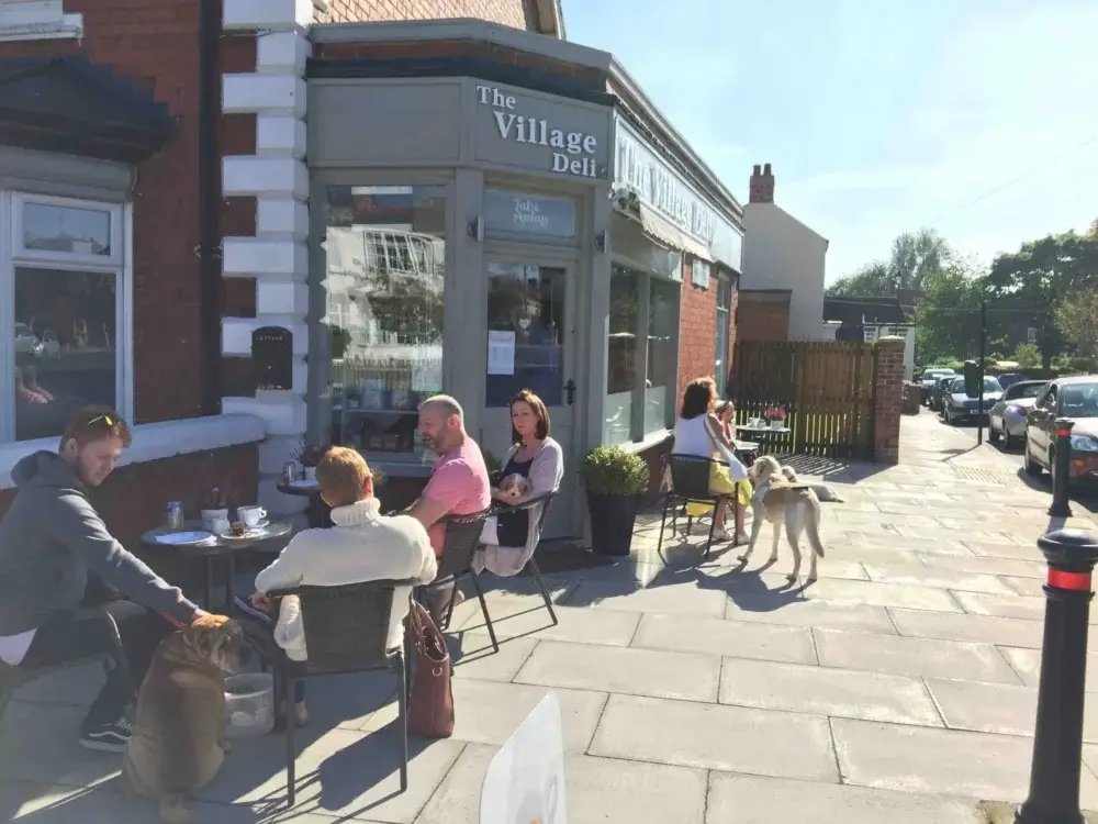 People sitting outside The Village Deli enjoying food and drinks, with dogs nearby on a sunny day. - Home Instead