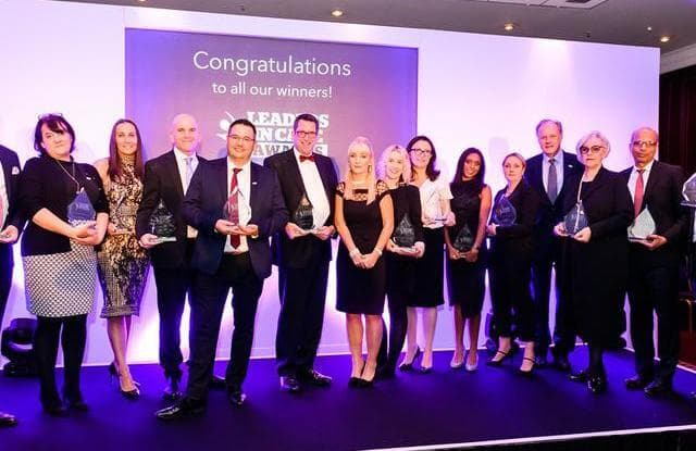 A group of people standing on stage holding awards with a "Congratulations to all our winners!" sign in the background. - Home Instead
