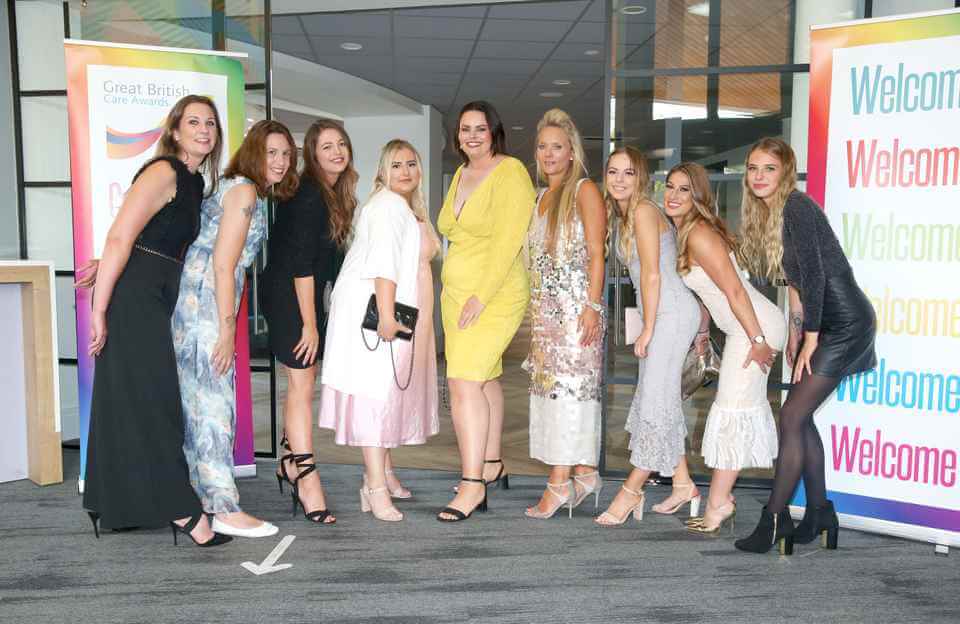 A group of women posing together in stylish outfits at an event with "Welcome" banners in the background. - Home Instead