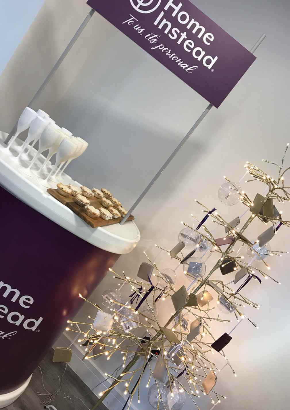 A decorated booth with a "Home Instead" sign, cookie tray, champagne glasses, and a lit tree display with hanging shapes. - Home Instead