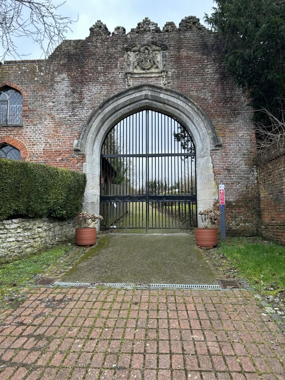 The entrance to Basing House, showing an old brick archway with a wrought iron gate, flanked by two potted plants, leading to a grassy area beyond.