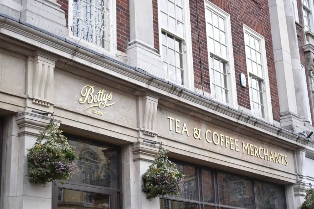 Storefront of Betty's Tea & Coffee Merchants with hanging flower baskets and large windows on a brick building. - Home Instead