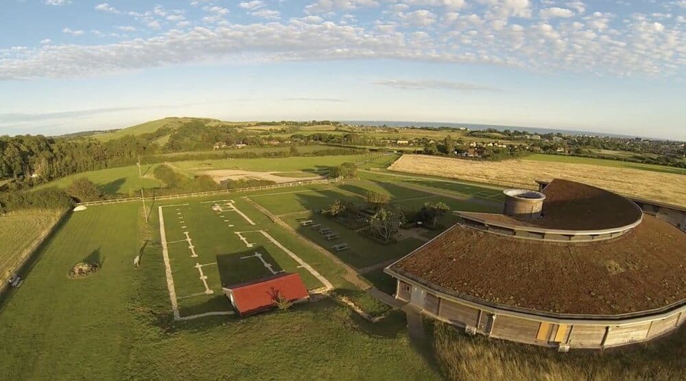 Aerial view of a lush green landscape with an ancient Roman ruin layout and an adjacent modern building under a partly cloudy sky. - Home Instead