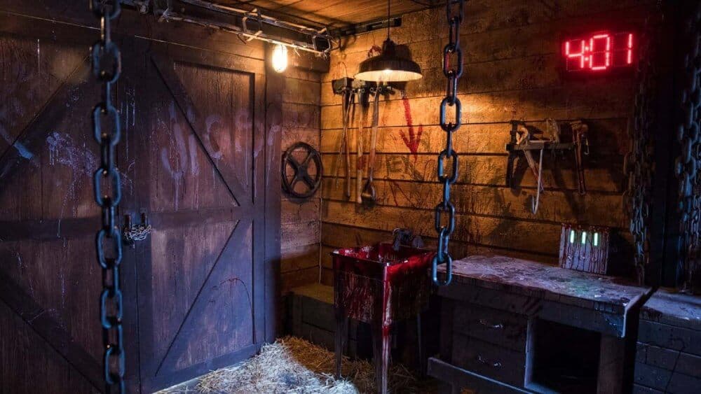 Dimly lit, rustic room with tools on walls, chains hanging, a red-lit timer reading 48:01, and blood-stained sink. - Home Instead