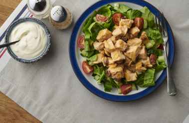 A plate of salad topped with grilled chicken pieces, next to a bowl of yogurt and salt and pepper shakers on a table. - Home Instead