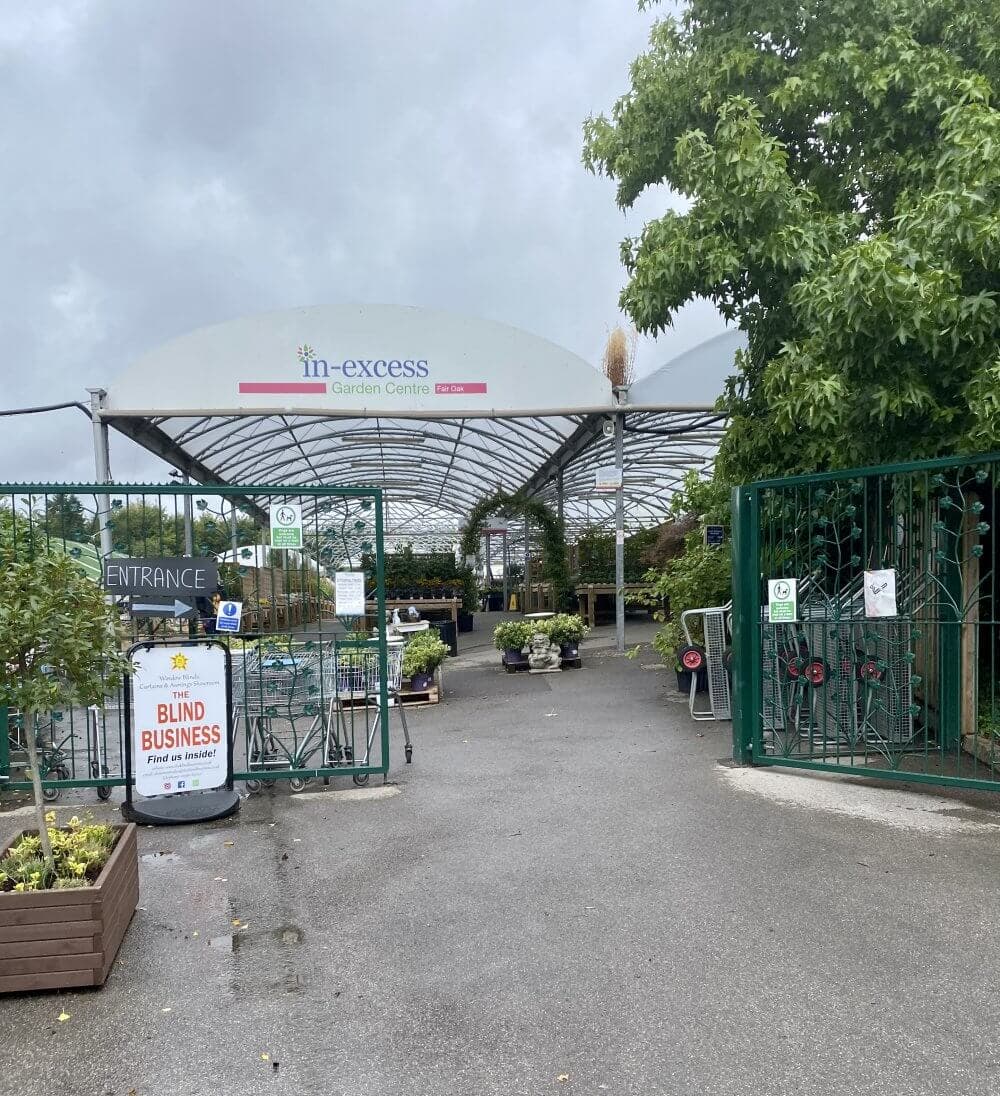 Entrance of a garden center with plants displayed, a large sign reading "In-Excess Garden Centre," and a "Blind Business" sign. - Home Instead