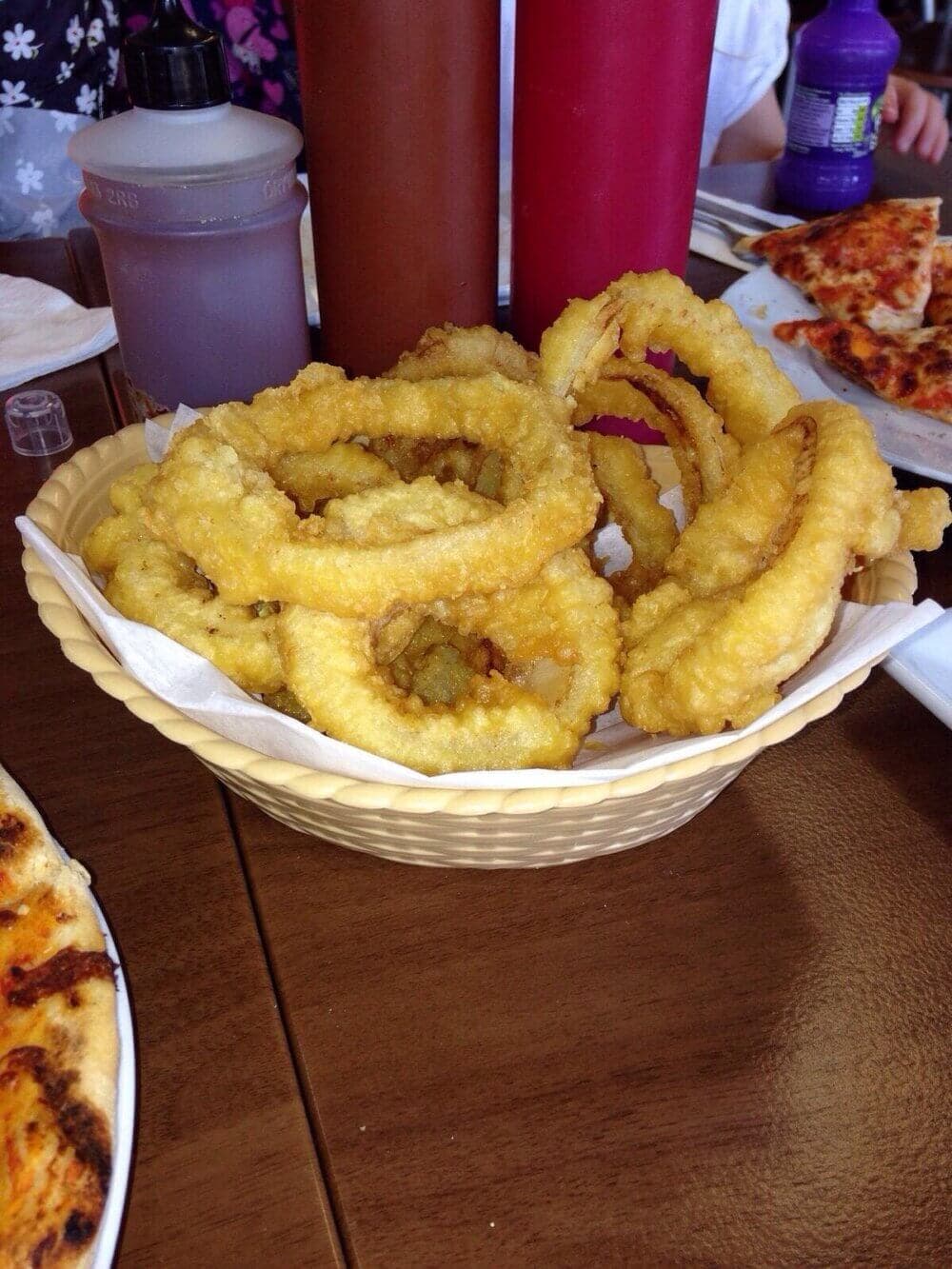 Dani's Diner - A basket of golden, crispy onion rings on a table with various condiments and pizza in the background. - Home Instead Renfrewshire and Barrhead care at home