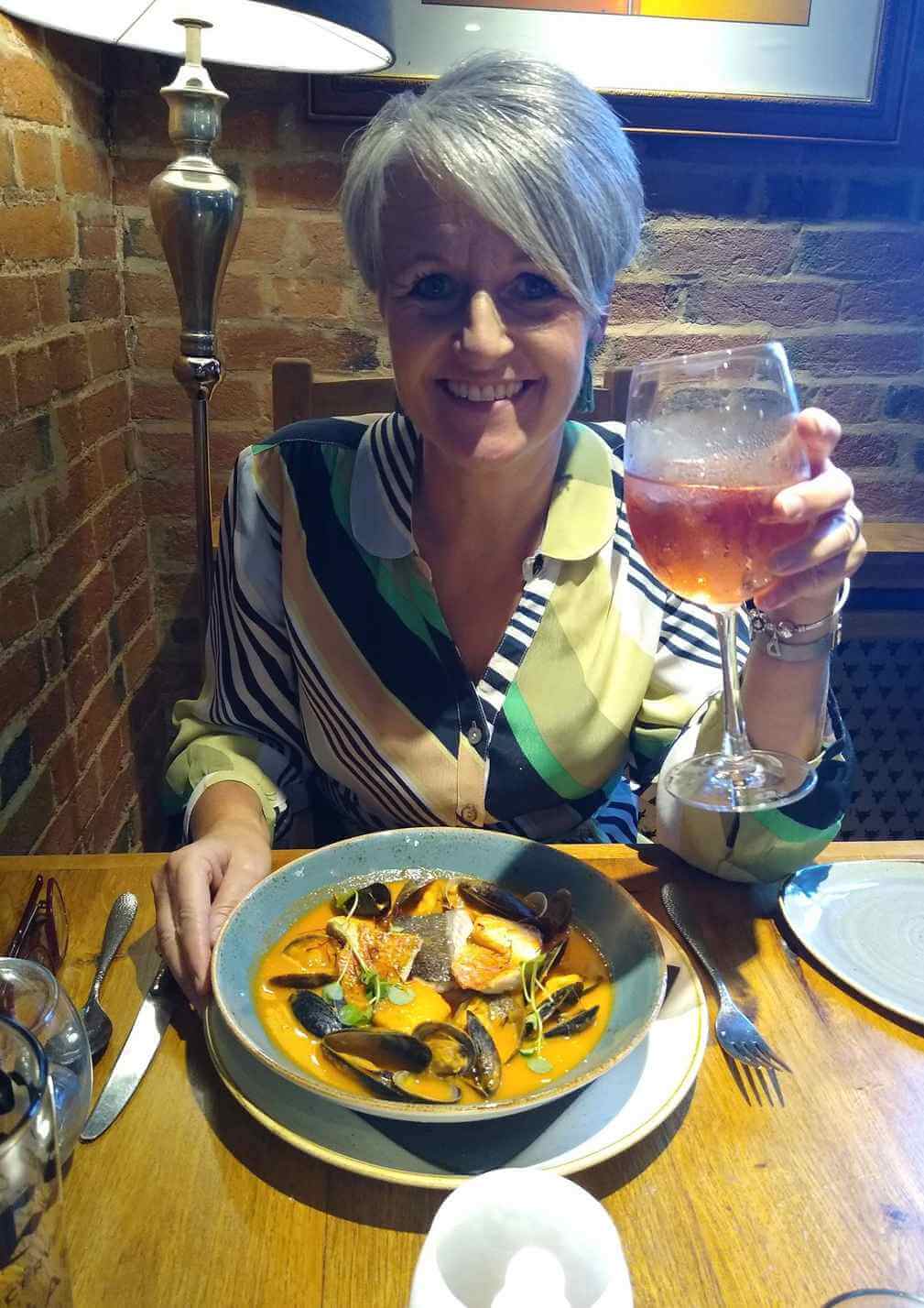 Person smiling, holding a glass of wine, and sitting at a table with a seafood dish in a brick-walled restaurant. - Home Instead