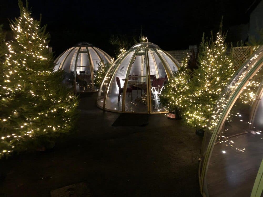 Three illuminated domes with string lights and Christmas trees at night. Inside each dome, a table and chairs are visible. - Home Instead