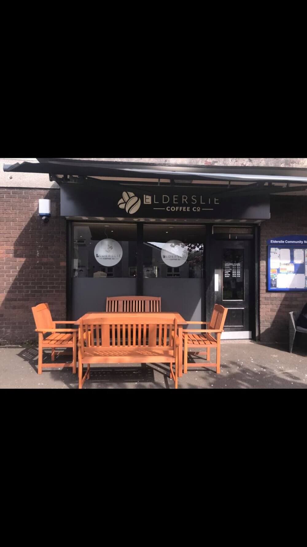 Outdoor seating area with wooden furniture in front of Elderslie Coffee Co. building with menu boards on either side. - Home Instead