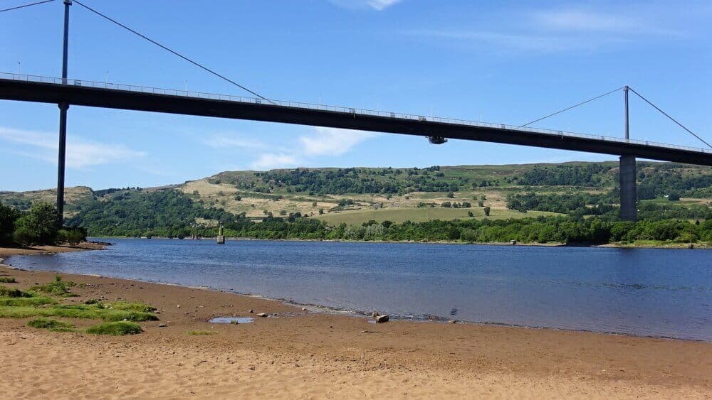 Erskine Bridge - A suspension bridge spans a wide river with grassy hills and a clear blue sky in the background. - Home Instead Renfrewshire and Barrhead homecare services
