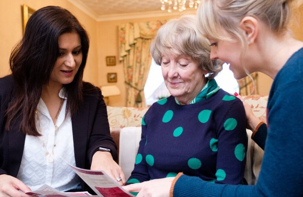 Three women sit together looking at papers, with one elderly woman in the middle and two younger women on each side. - Home Instead