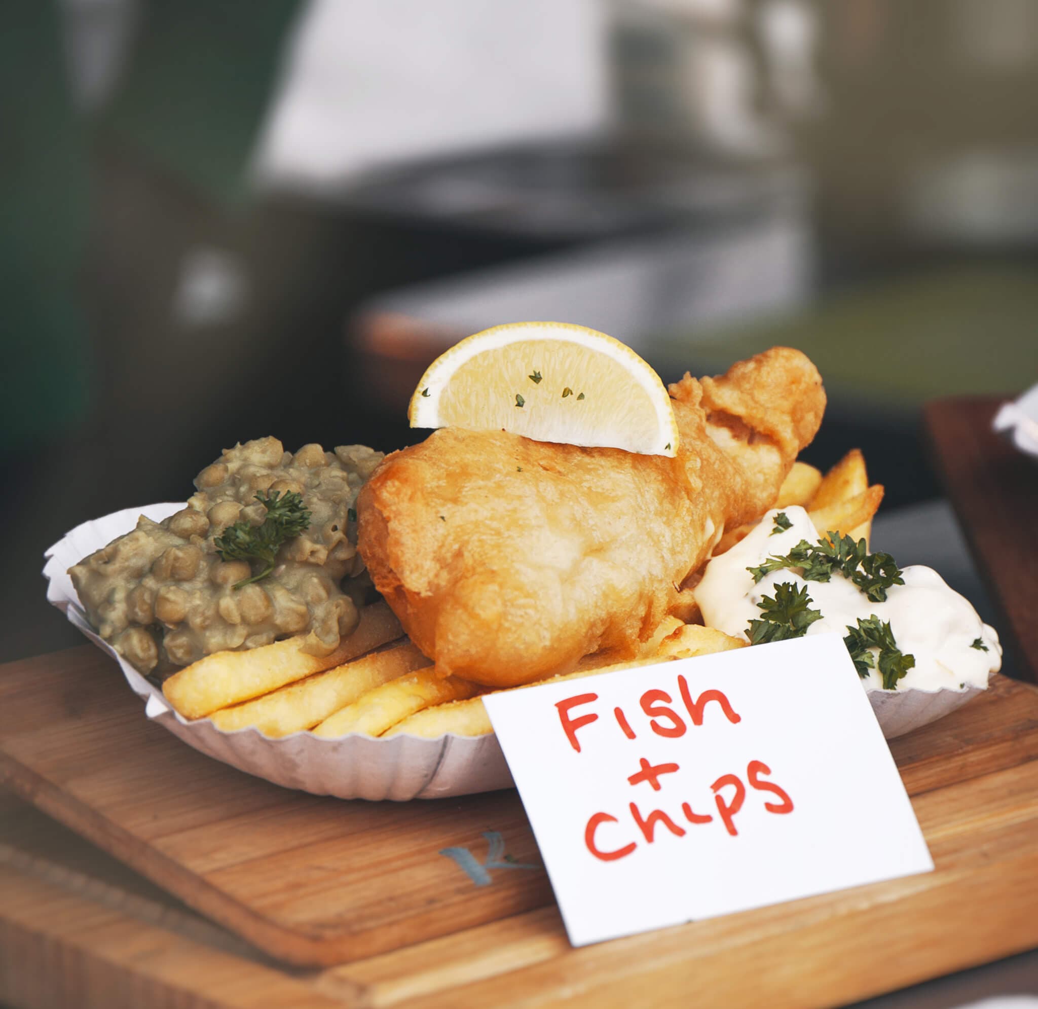 A dish of fish and chips with mushy peas and tartar sauce on a wooden board, garnished with lemon and parsley. - Home Instead Renfrewshire and Barrhead care at home services