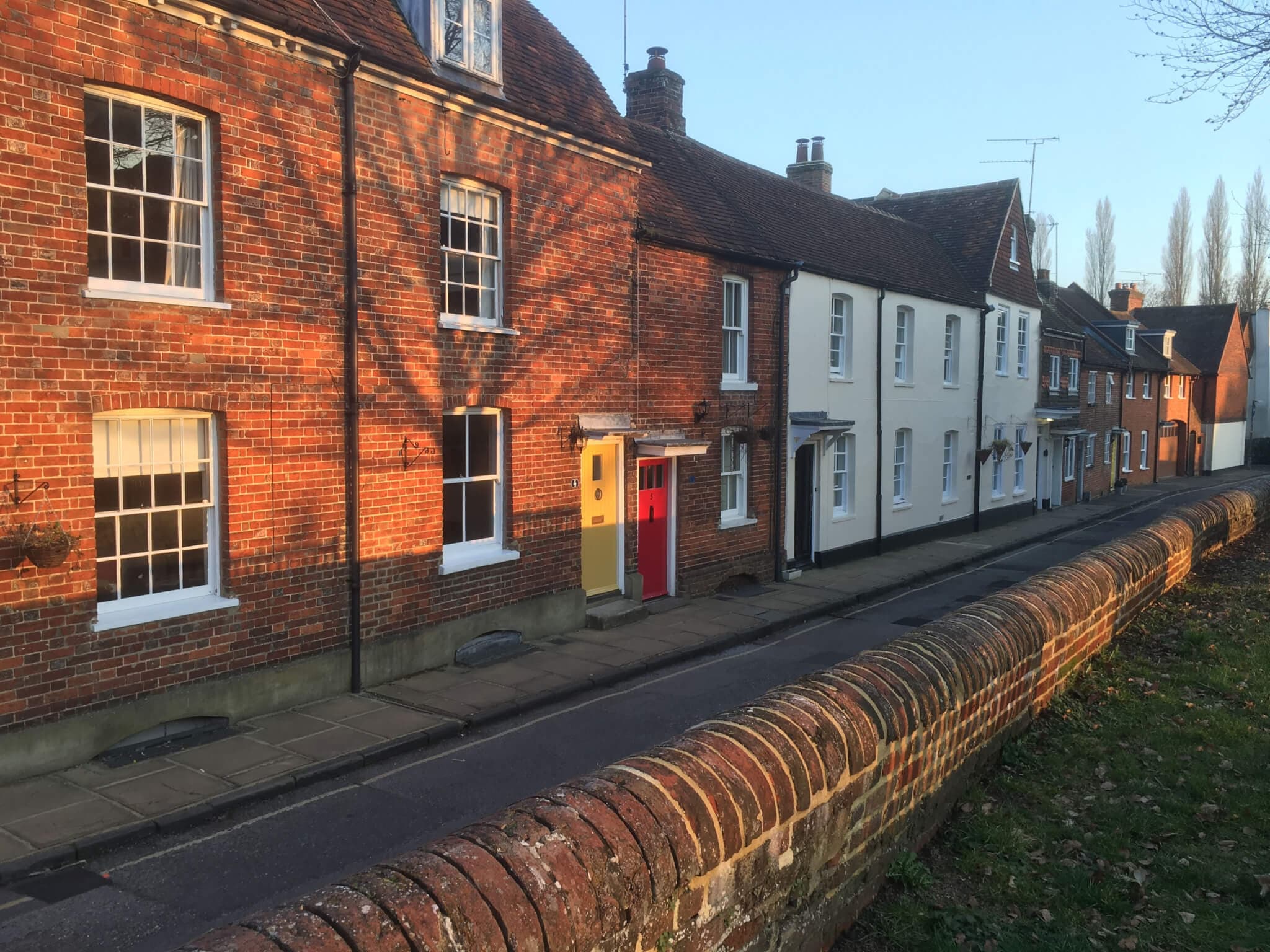 A row of English terraced houses with red and yellow doors, lit by the setting sun, and a brick wall in the foreground. - Home Instead