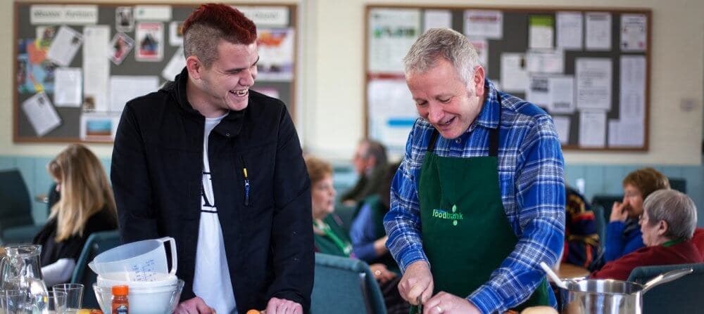 Two men smiling and cooking together. One wears a black jacket, and the other a blue plaid shirt and a green apron. - Home Instead